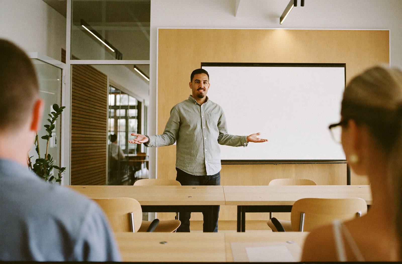 Un maestro da una clase muestra frente a un pizarrón blanco en un salón moderno; dos personas de la escuela lo observan.