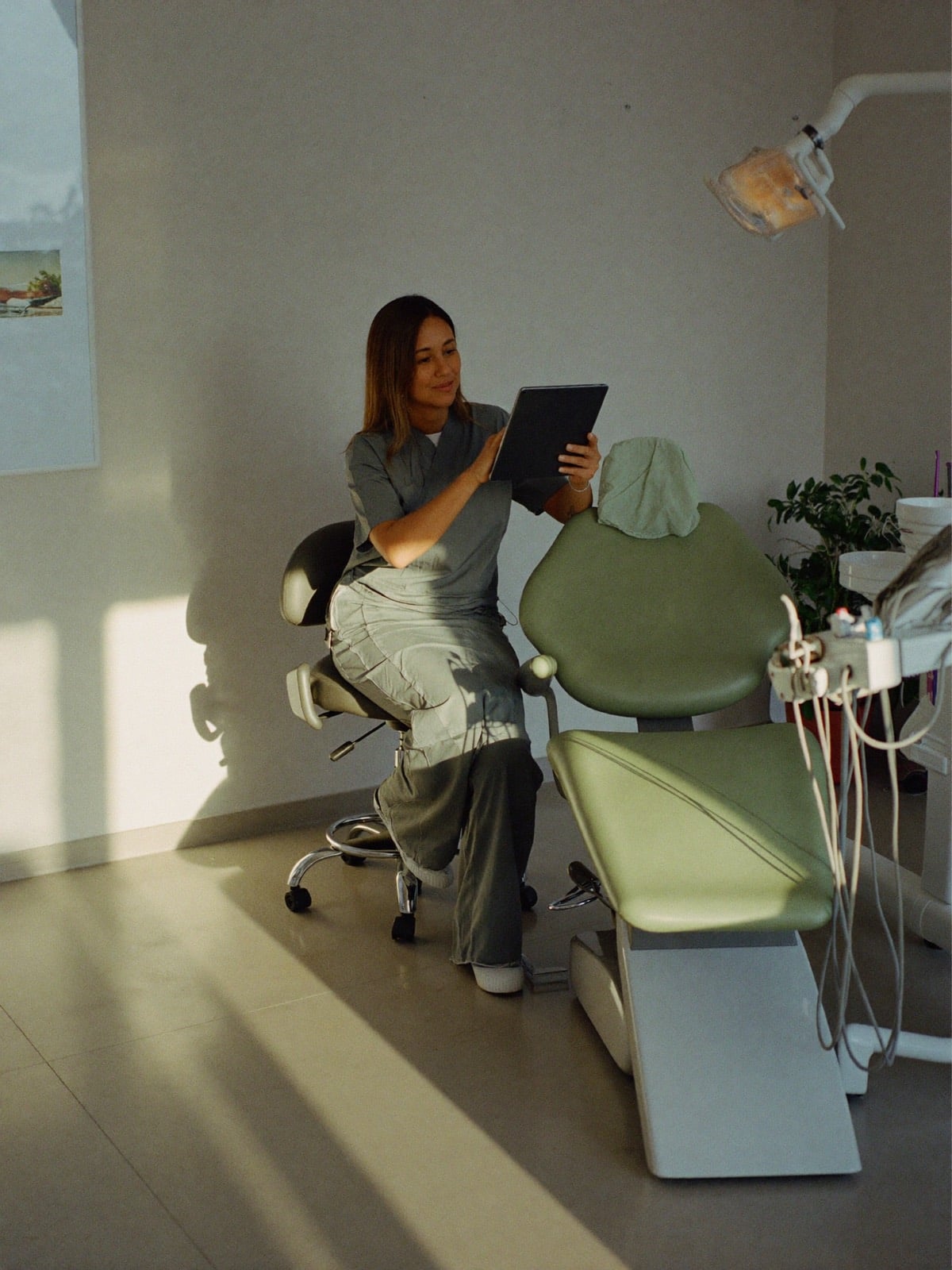 Una dentista revisa una tableta sentada junto al sillón de su consultorio, con la luz de la tarde.