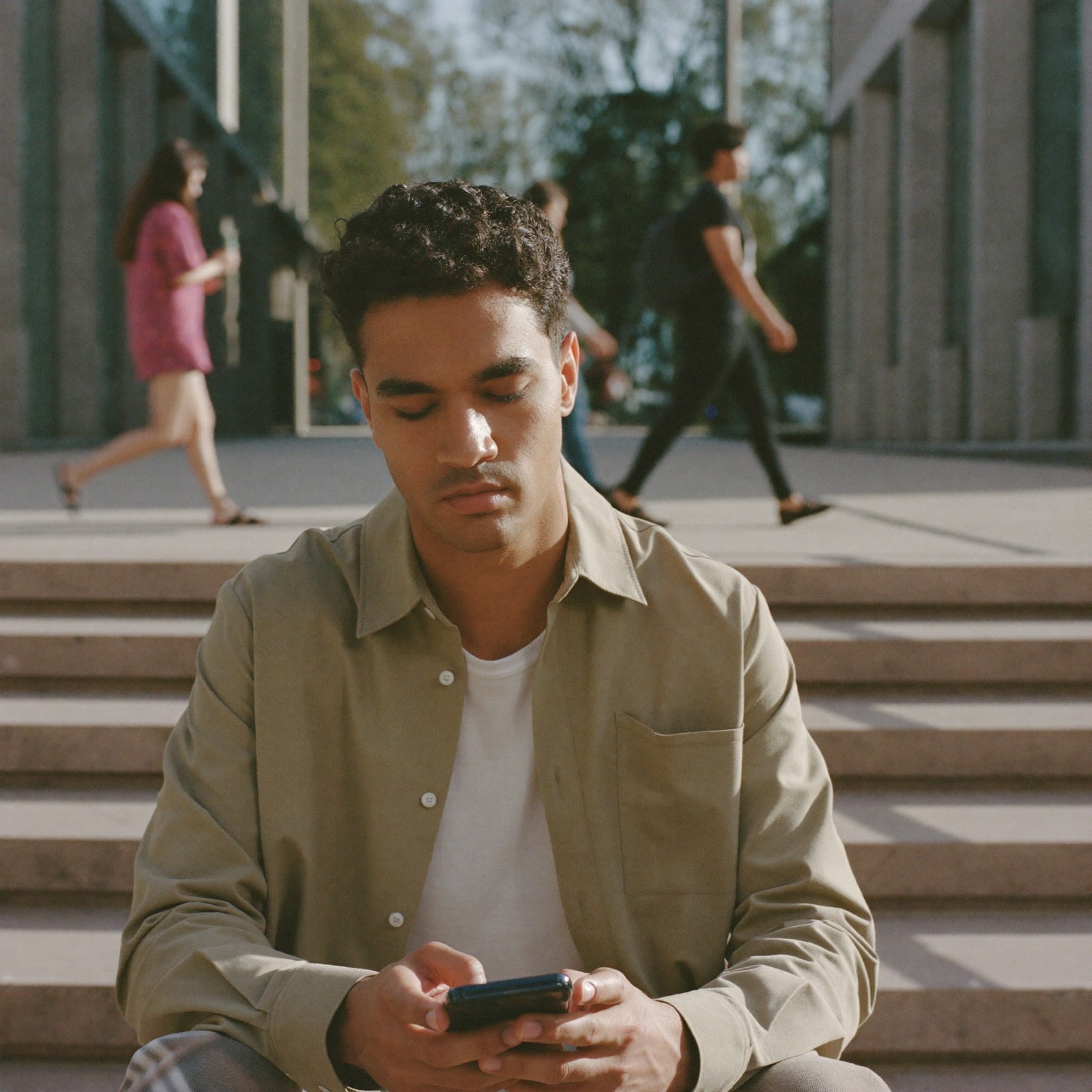 Un joven contesta en su celular sentado en unas escaleras, con gente que pasa detrás.